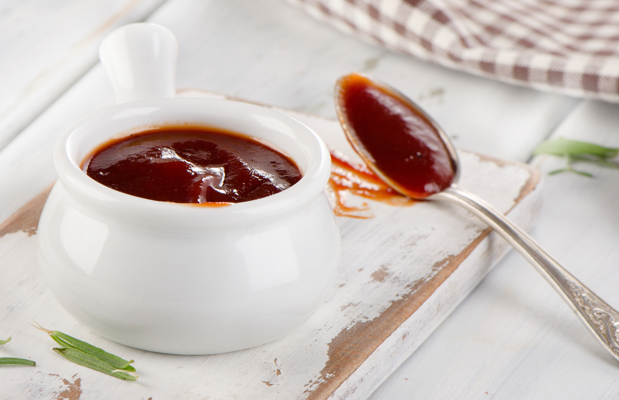 Barbeque sauce in a white dish on a white plate with a spoon and napkin pictured on a table