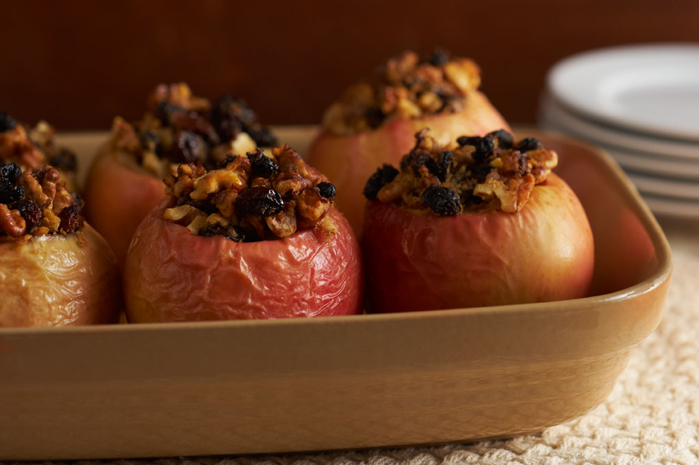 Baked red apples stuffed with oats in a baking pan with plates in the background on a table