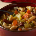 Bean and vegetable soup in a red bowl on a white tablecloth with a spoon