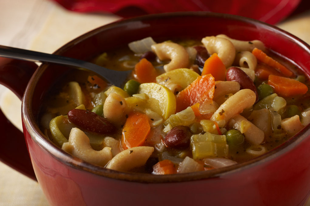 Bean and vegetable soup in a red bowl on a white tablecloth with a spoon