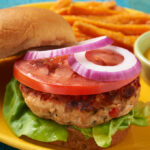 Chicken burger on a yellow plate pictured on a blue tablecloth with mustard, tomato and onion.