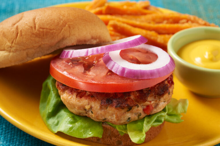 Chicken burger on a yellow plate pictured on a blue tablecloth with mustard, tomato and onion.