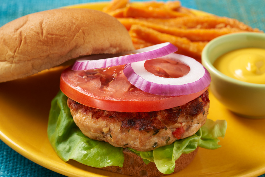 Chicken burger on a yellow plate pictured on a blue tablecloth with mustard, tomato and onion.