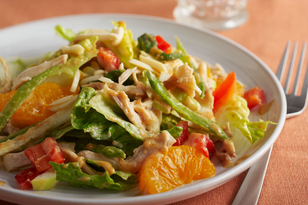 Chicken salad with peanut dressing on a white plate pictured on a wooden table with a fork and glass.