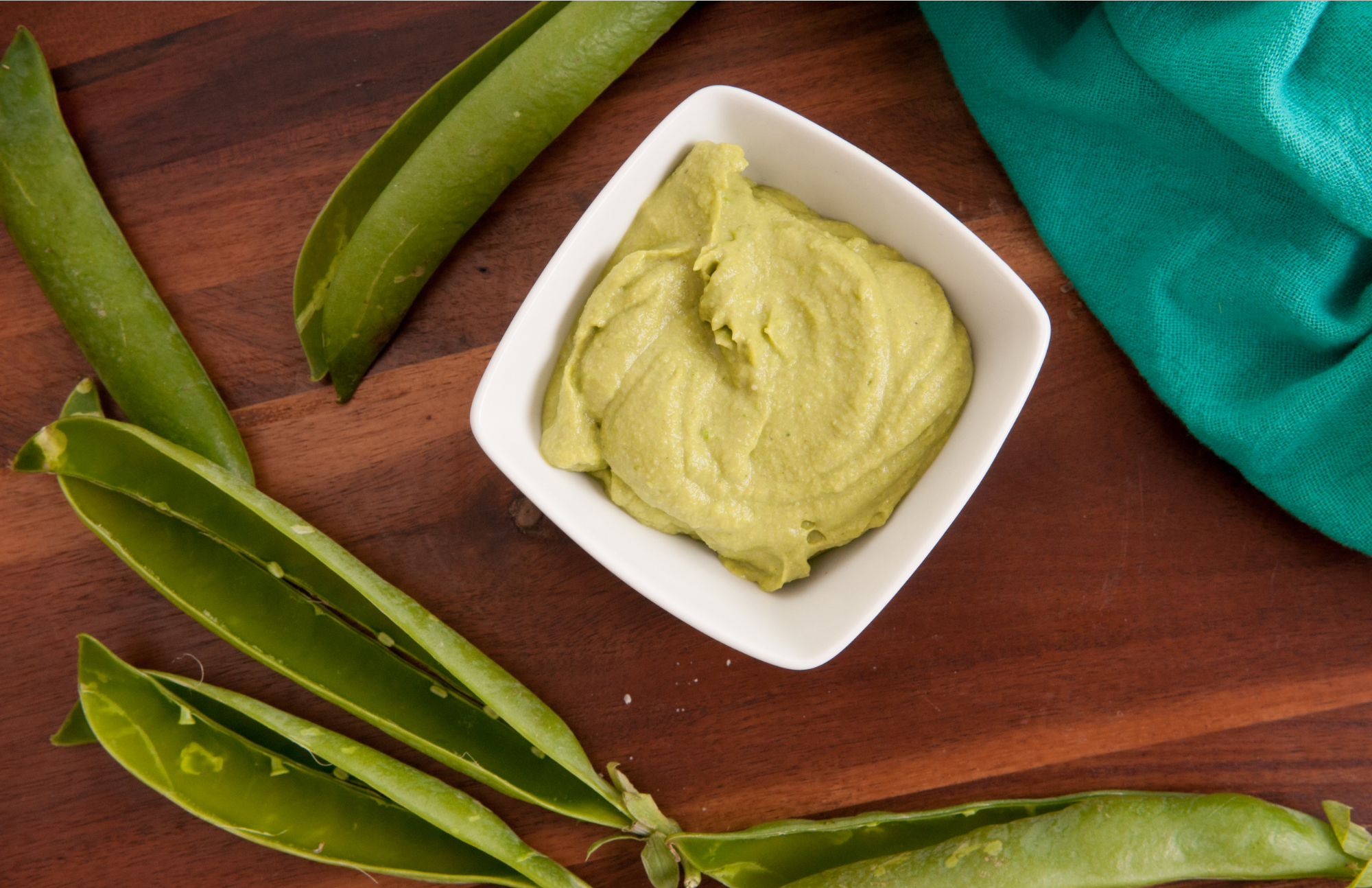 White bowl on a wooden table with green napkin close by. The bowl is filled with pea pesto and is surrounded by snap pea shells.