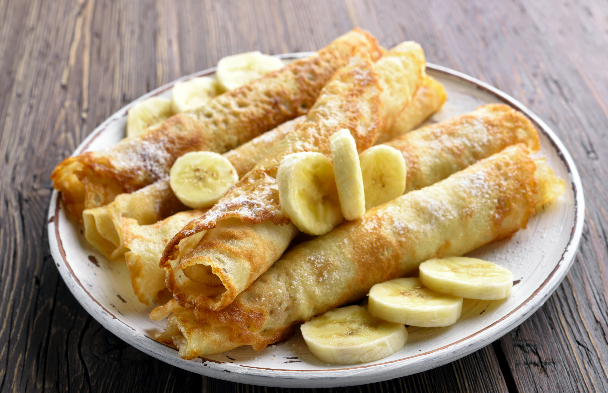 Whole wheat crepes with syrup and bananas pictured on a white plate on a wooden table
