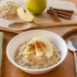 Apple Cinnamon Oatmeal in a bowl with spoon and cutting board with apple and cinnamon sticks in the background