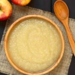 Applesauce in a wooden bowl with wooden spoon and two apples in the background on a black table