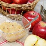 Applesauce with red apples on a a red and white tablecloth with apples in a basket.