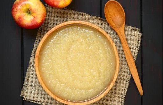 Applesauce in a wooden bowl with wooden spoon and two apples in the background on a black table