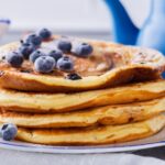 Blueberry pancakes on a white and blue plate with a blue pitcher.