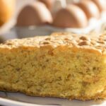 Carrot cake slice on a white plate with eggs in the background.