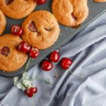 Cherry muffins in a tin on a grey table with cherries in the front