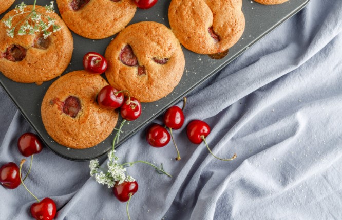 Cherry muffins in a tin on a grey table with cherries in the front