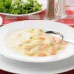 Chicken and dumplings in a white bowl with spoon on red check tablecloth