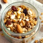 Homemade granola in a glass jar on a white napkin with nuts, oats and a spoon.