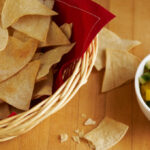 Homemade corn tortilla chips in a basket lined with red napkins pictured on a wooden table.