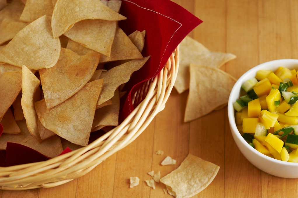 Homemade corn tortilla chips in a basket lined with red napkins pictured on a wooden table.