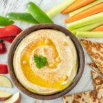 Hummus in a brown bowl surrounded by fresh fruit and vegetables.