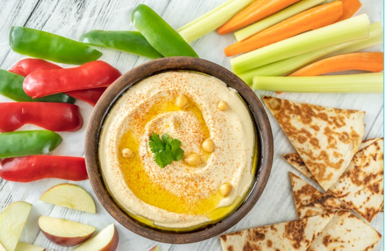 Hummus in a brown bowl surrounded by fresh fruit and vegetables.