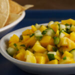 Mango salsa in a white bowl on top of a blue tablecloth with a chips in a white bowl.