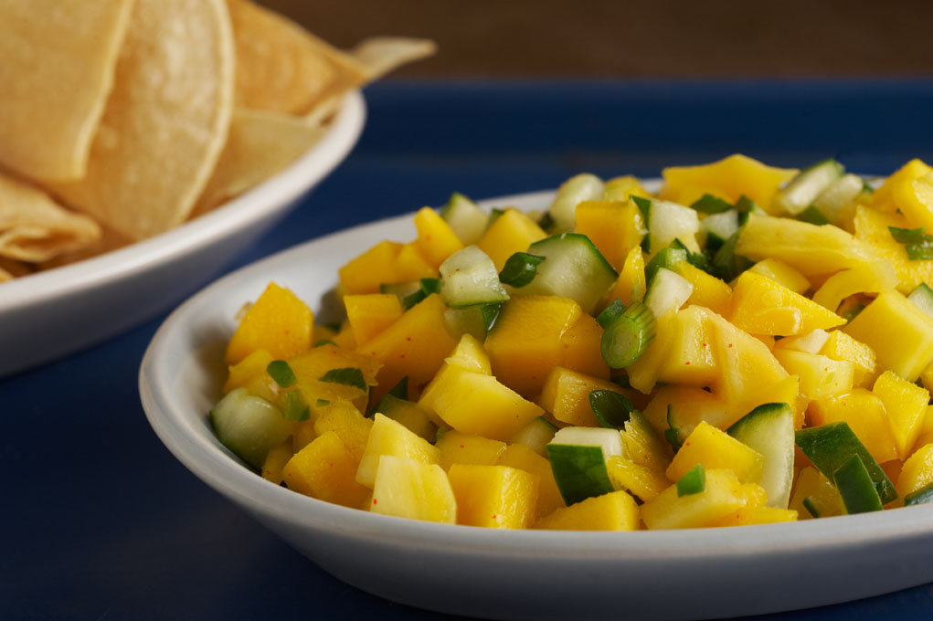 Mango salsa in a white bowl on top of a blue tablecloth with a chips in a white bowl.