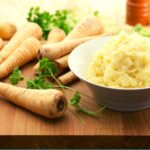 Mashed parsnips in a white bowl next to fresh parsnips on a wooden table.
