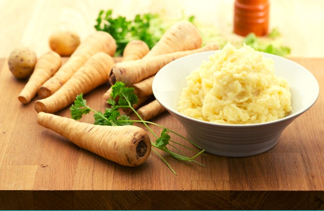 Mashed parsnips in a white bowl next to fresh parsnips on a wooden table.