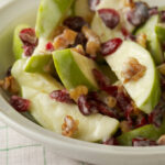 A walnut, apple, and cranberry salad in a white bowl atop a green and white tea towel