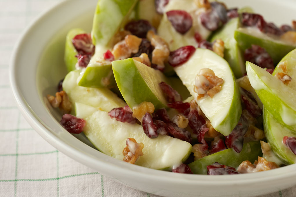 A walnut, apple, and cranberry salad in a white bowl atop a green and white tea towel