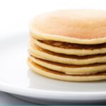 A stack of pancakes on a white plate on top of a white table.