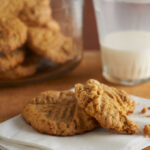 Rolled oat and peanut butter cookies on a white napkin with a jar of cookies and a half full glass of milk in the background