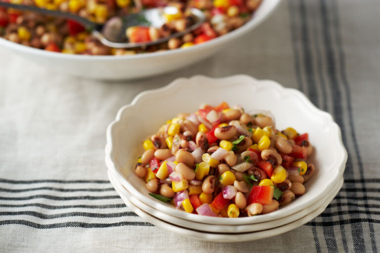 White bowl filled with southwestern black-eyed pea and corn salad with a white serving serving bowl in the background.