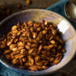 Spicy pumpkin seeds in a grey bowl on blue cloth with a metal spoon.