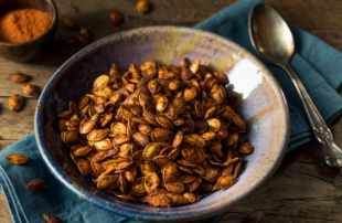 Spicy pumpkin seeds in a grey bowl on blue cloth with a metal spoon.