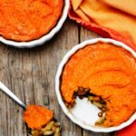 Two white pie dishes containing sweet potato shepherd's pies atop a wooden surface. A spoonful of pie and orange fabric decoratively surround the pie dishes.