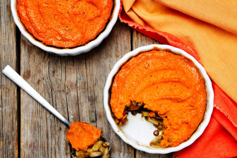 Two white pie dishes containing sweet potato shepherd's pies atop a wooden surface. A spoonful of pie and orange fabric decoratively surround the pie dishes.