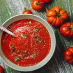 Tomato soup in a white bowl on a green wooden table with fresh tomatoes.