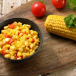 Black bowl with tomato and corn with corn on the cob, tomatoes, and green herbs on the side.