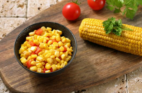 Black bowl with tomato and corn with corn on the cob, tomatoes, and green herbs on the side.