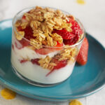 Clear cup with yogurt, fruit, and granola on top of a blue plate.