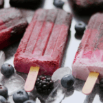 Three berry good popsicles with blueberries and blackberries scattered around the popsicles.