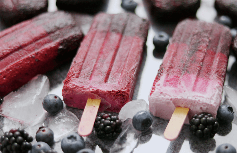 Three berry good popsicles with blueberries and blackberries scattered around the popsicles.