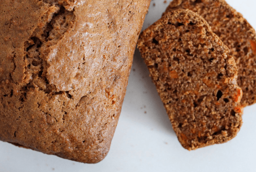 Two slices of carrot bread next to a carrot bread loaf on a white surface.