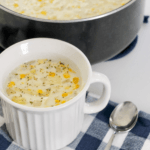 A white cup with corn chowder on top of a blue and white checkered cloth.