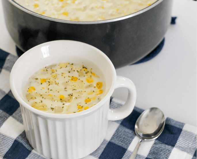 A white cup with corn chowder on top of a blue and white checkered cloth.