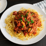 Red lentil bolognese on top of spaghetti noodles on a white plate.