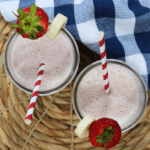 Two glasses of oatmeal, strawberry, banana smoothies garnished with a strawberry and banana with a red- and white-striped straw.