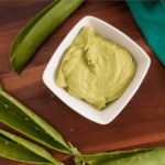 Pea pesto in a white bowl surrounded by empty pea pods