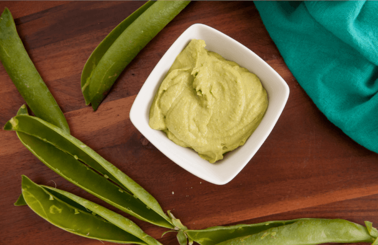 Pea pesto in a white bowl surrounded by empty pea pods